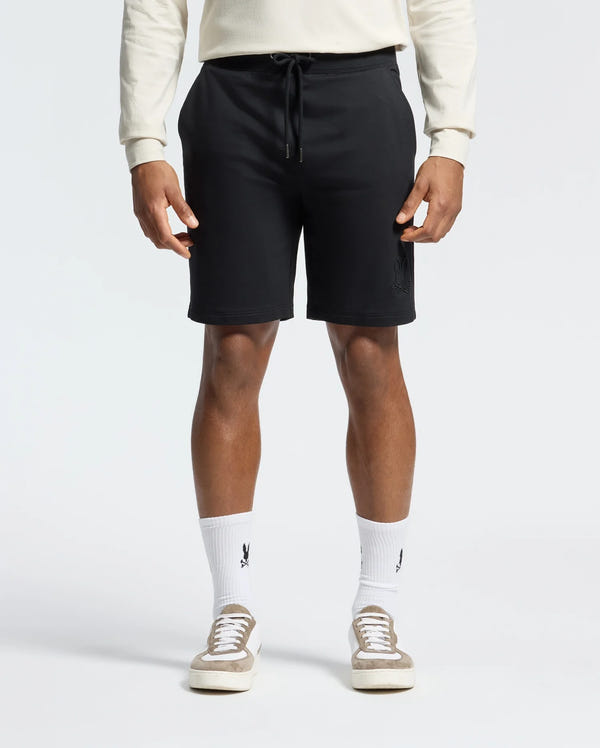 A man poses against a white background, wearing a cream long-sleeve shirt and Psycho Bunny's MENS LEVANZO EMBROIDERED MICRO FRENCH TERRY SWEATSHORT in black for ultimate comfort. His look is finished with white logo socks and beige sneakers.