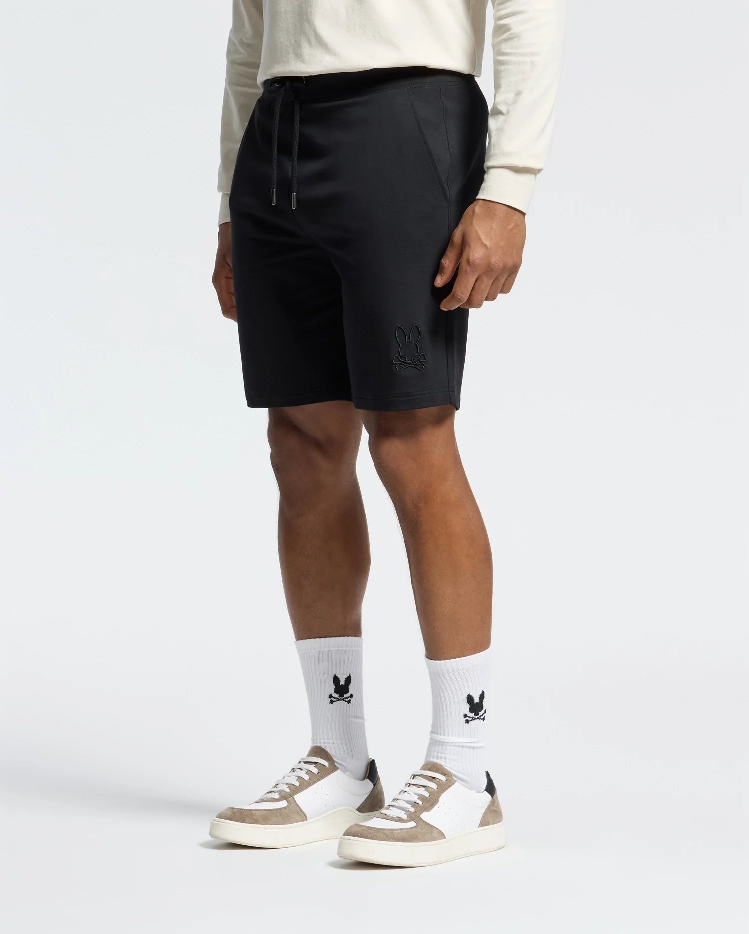 A person wears a long-sleeve shirt, Psycho Bunny's Levanzo Embroidered Micro French Terry Sweatshorts (B6R578F200) with an Embroidery Bunny emblem, motif-matching white socks, and white sneakers with beige accents. The sporty outfit contrasts a plain white backdrop.