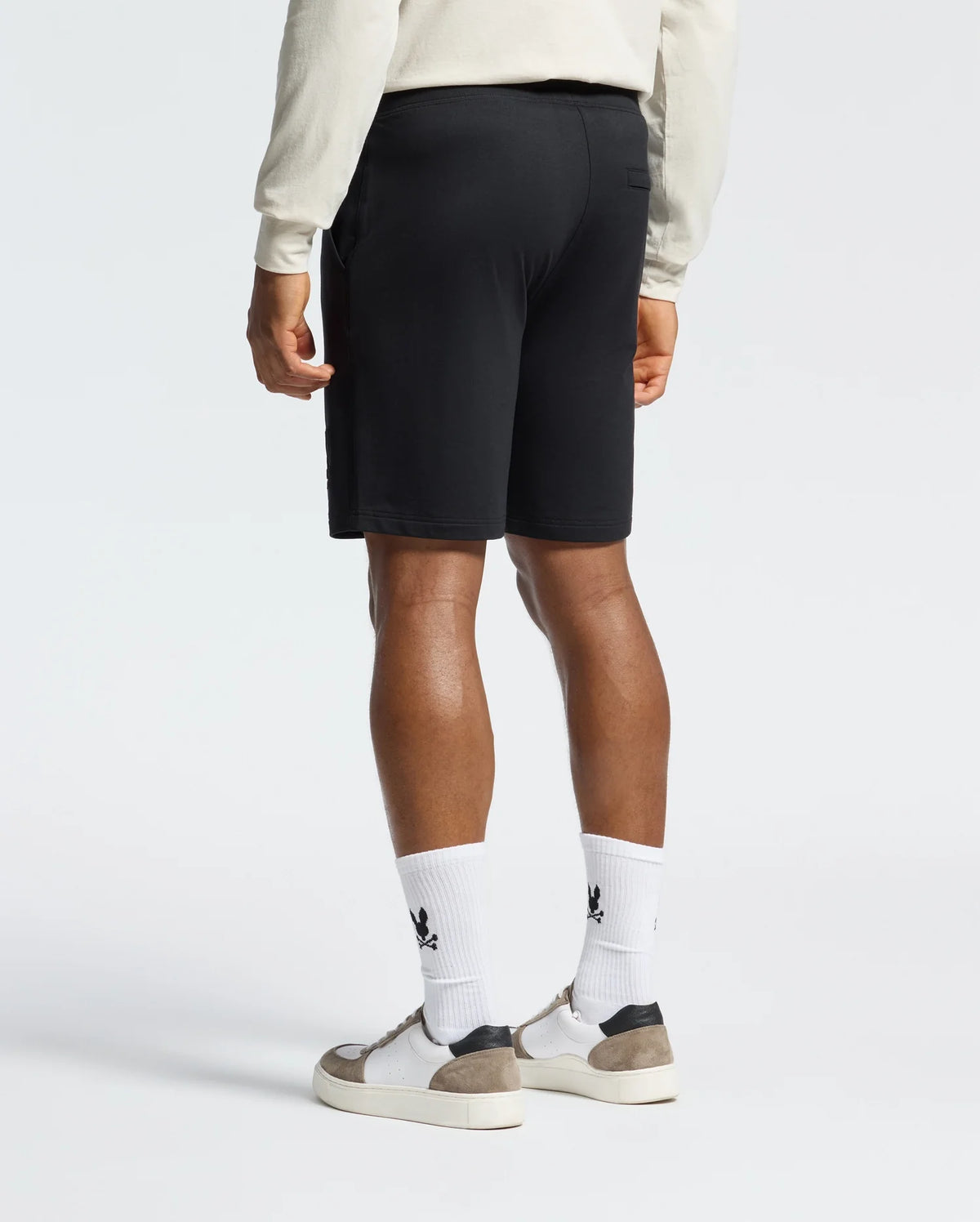 A person is wearing a cream long-sleeve shirt, Psycho Bunny's Levanzo Embroidered Micro French Terry Sweatshorts, and white socks with black logos, paired with white and gray sneakers. The Embroidery Bunny on the shorts adds charm as they stand against a plain background, shown from behind.