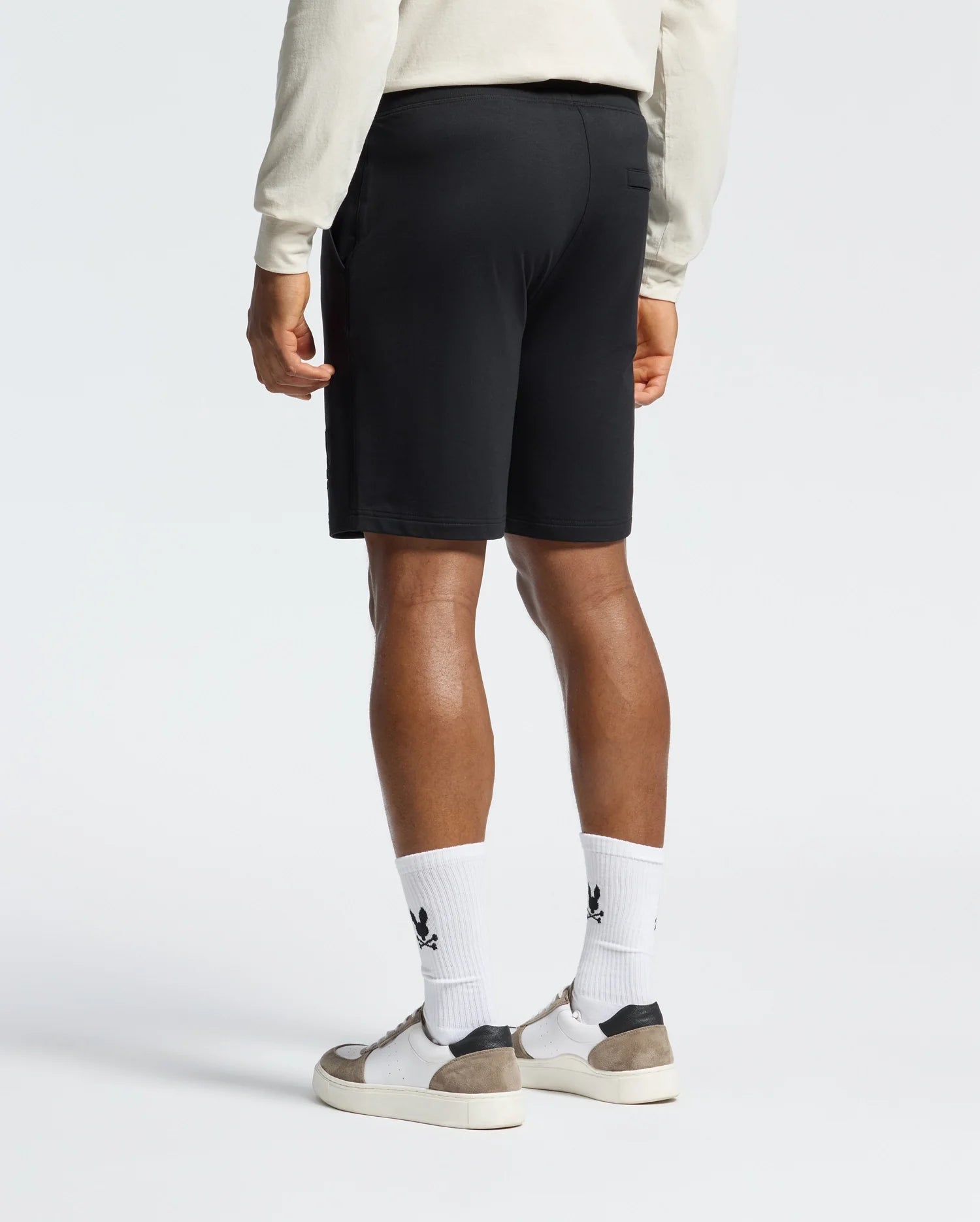 A person is wearing a cream long-sleeve shirt, Psycho Bunny's Levanzo Embroidered Micro French Terry Sweatshorts, and white socks with black logos, paired with white and gray sneakers. The Embroidery Bunny on the shorts adds charm as they stand against a plain background, shown from behind.