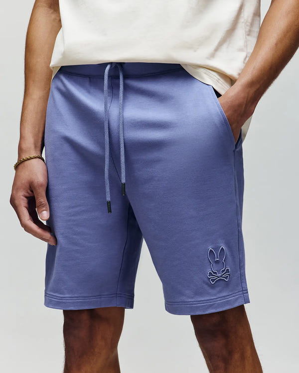 A person wears Psycho Bunny’s MENS LEVANZO EMBROIDERED MICRO FRENCH TERRY SWEATSHORT (B6R578F200) in light purple, featuring an embroidered Bunny and crossbones logo on the left leg, paired with a cream t-shirt and hands in pockets.