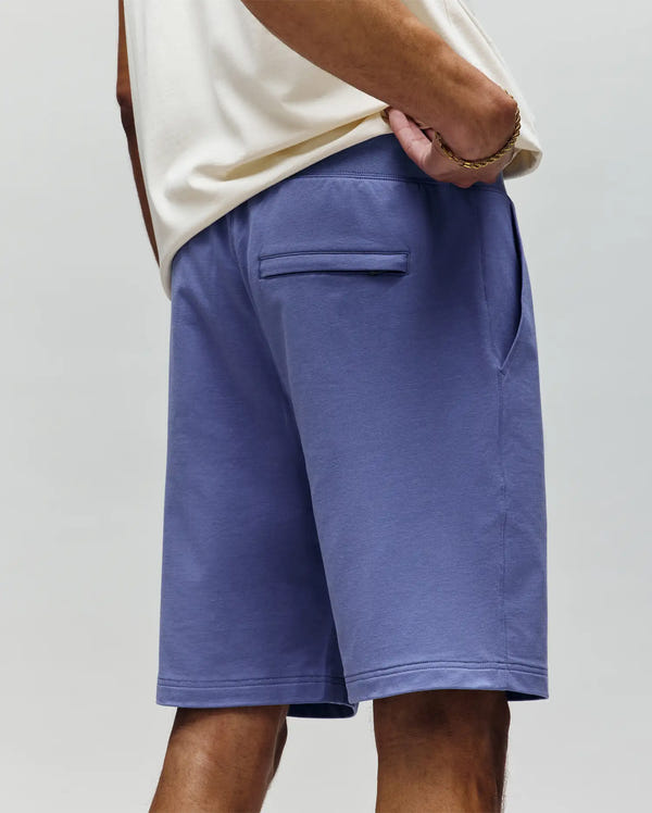 A person stands with one hand in their pocket, wearing Psycho Bunny MENS LEVANZO EMBROIDERED MICRO FRENCH TERRY SWEATSHORT - B6R578F200 in light purple and a cream shirt, highlighting the shorts’ rear pocket and relaxed fit from the back and side view.