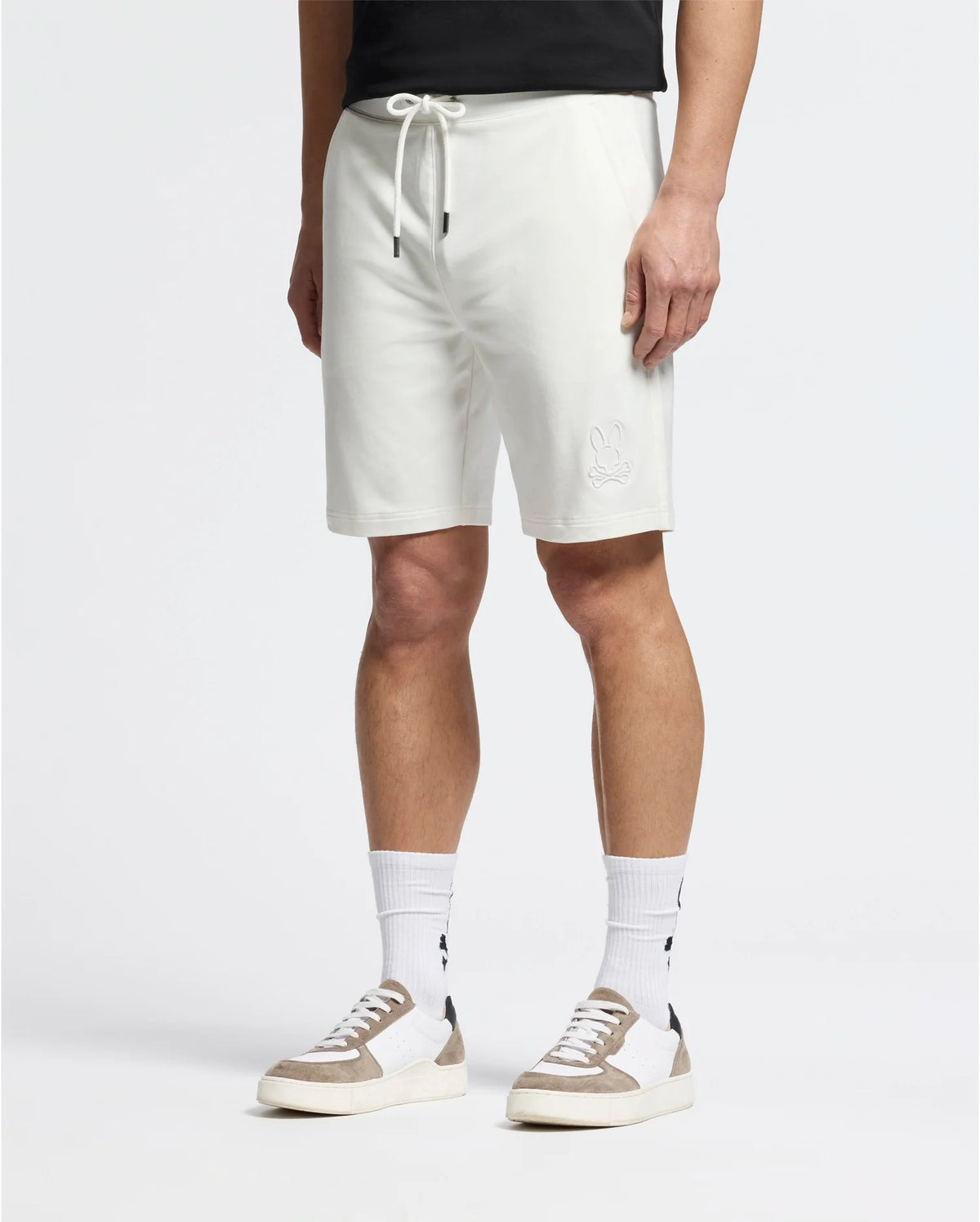 A person in Psycho Bunny MENS LEVANZO EMBROIDERED MICRO FRENCH TERRY SWEATSHORT (B6R578F200), a black t-shirt, white crew socks with black designs, and white sneakers with beige accents stands against a plain background.