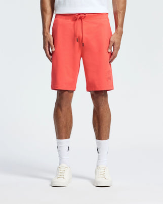 Wearing coral Psycho Bunny Levanzo sweatshorts made from micro French terry, a person pairs them with a white t-shirt, coordinating crew socks, and sleek sneakers. The shorts feature an embroidered Bunny logo on the left leg, set against a plain white background that highlights the vibrant outfit.