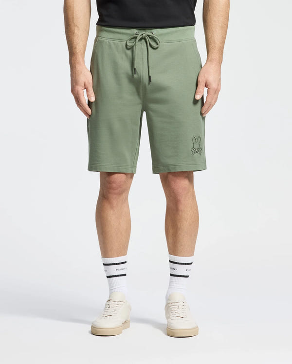 A person is wearing sage green Psycho Bunny Men's Levanzo Embroidered Micro French Terry Sweatshorts (B6R578F200) with a black top, white socks featuring an embroidered bunny logo, and white sneakers against a plain white background.