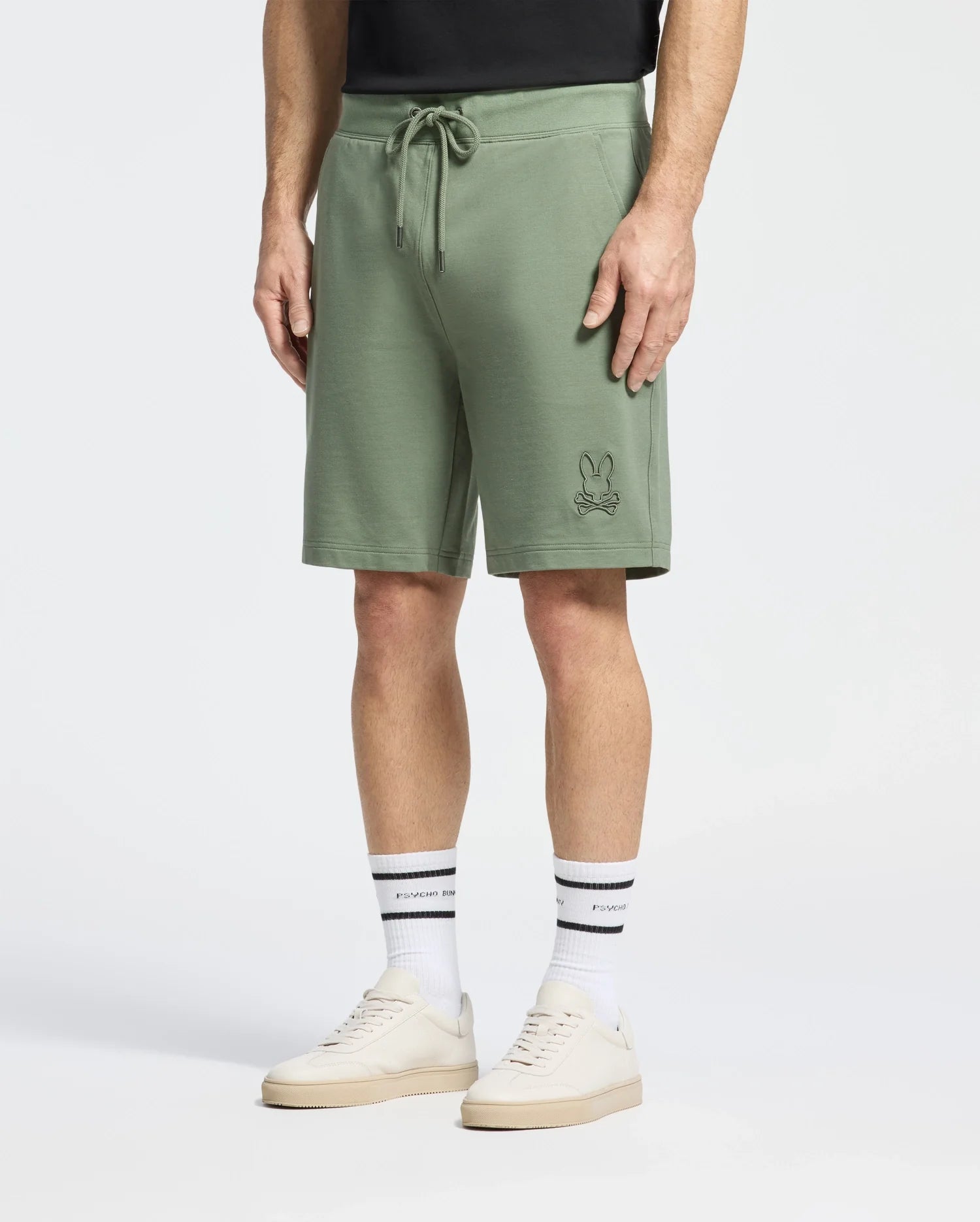 A person wears Psycho Bunny's Levanzo Embroidered Micro French Terry Sweatshorts with a bunny logo, paired with a black shirt, white socks with black stripes and text, and light beige sneakers against a light gray backdrop.