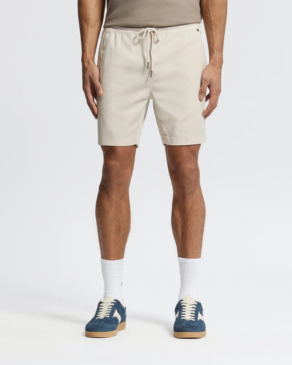 Effortlessly stylish, a person wears beige Psycho Bunny MENS WILLIS STRETCH TENCEL SHORTS (B6R584F200) with an elastic waistband, a taupe tank top, white socks, and blue sneakers against a plain white background.