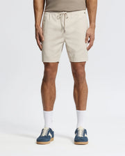 Effortlessly stylish, a person wears beige Psycho Bunny MENS WILLIS STRETCH TENCEL SHORTS (B6R584F200) with an elastic waistband, a taupe tank top, white socks, and blue sneakers against a plain white background.