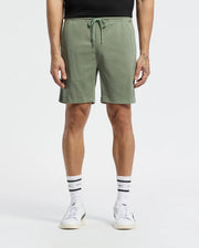 A person wears Psycho Bunny MENS WILLIS STRETCH TENCEL SHORT - B6R584F200 in sage green, paired with a black shirt, white socks with black stripes, and white sneakers. Only the lower torso and legs are visible against a plain white background.