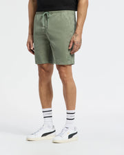 A person wearing Psycho Bunny MENS WILLIS STRETCH TENCEL SHORT in sage green (style B6R584F200), a black shirt, white socks with black stripes, and white sneakers with black accents, stands against a plain light background.
