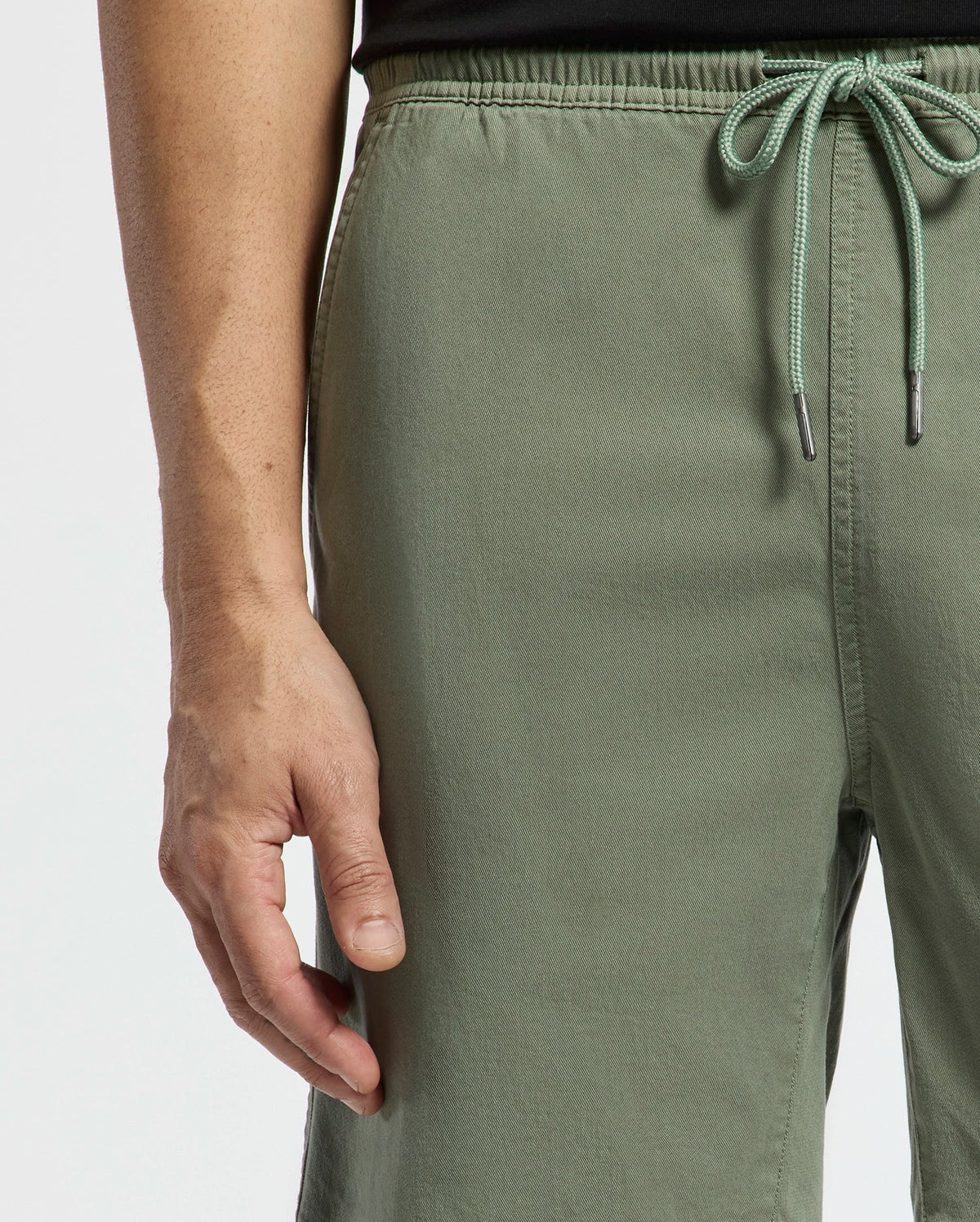 A close-up shows a person’s lower torso and hand, wearing Psycho Bunny MENS WILLIS STRETCH TENCEL SHORT - B6R584F200 in green and a black top, with their hand relaxed at their side against a plain background.