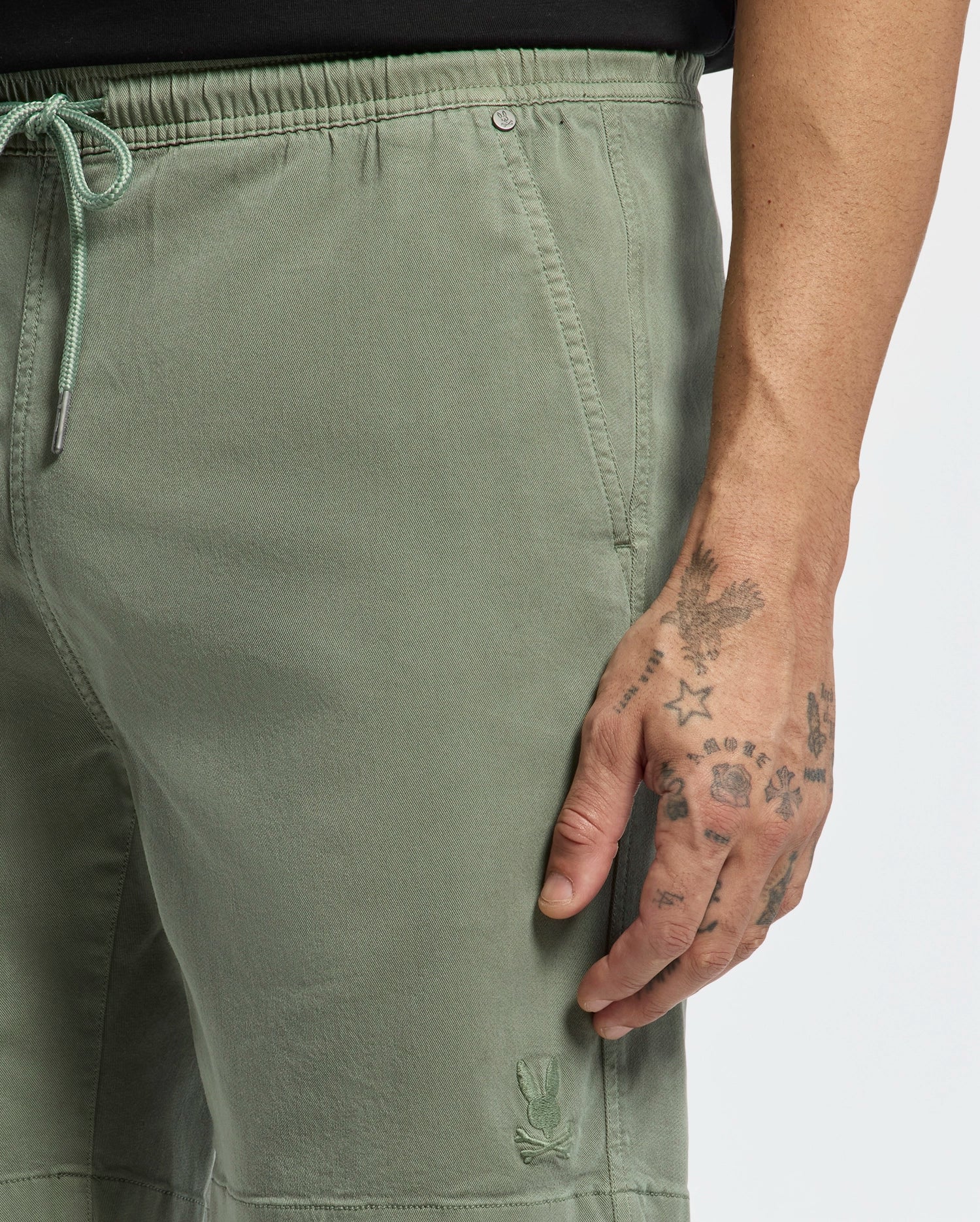 A person wears Psycho Bunny MENS WILLIS STRETCH TENCEL SHORT - B6R584F200 in sage green, featuring a drawstring waist, visible stitching, and a small embroidered bunny near the hem; their tattooed right hand rests by their side.