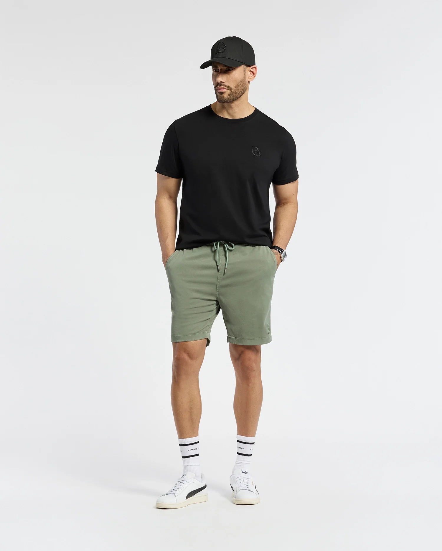 A man in a Psycho Bunny regular fit black t-shirt, MENS WILLIS STRETCH TENCEL SHORT - B6R584F200 green shorts, and white sneakers stands against a white background, hands in pockets. He sports an embroidered Bunny cap and stretch Tencel™ blend socks with black stripes.