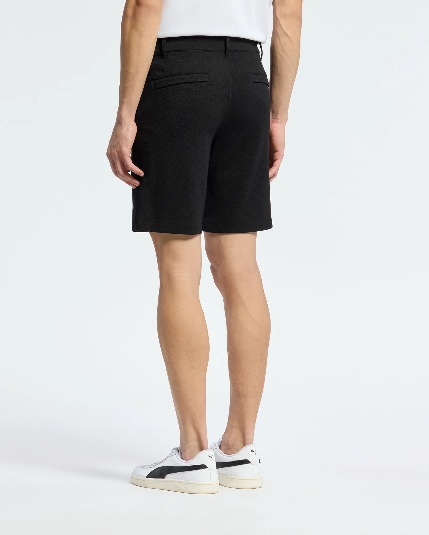A person stands with their back to the camera, wearing a white shirt and the Psycho Bunny MENS SHIRO KNIT HONEYCOMB SHORT - B6R585F200. White sneakers with black accents complete the look against a plain background, showcasing a stylish transition for any setting.