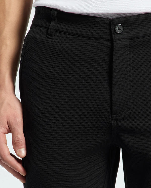 A close-up shows someone wearing Psycho Bunny's MENS SHIRO KNIT HONEYCOMB SHORT in black, with their hand resting near the waistband. The stylish honeycomb pattern and a black button enhance the sophisticated look of this chic ensemble, complemented by a neatly tucked white shirt.