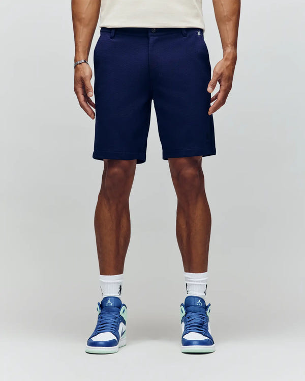 A person wearing Psycho Bunny's MENS SHIRO KNIT HONEYCOMB SHORT - B6R585F200 in navy blue, paired with white logo socks and blue and white sneakers, stands against a plain light gray background; only their lower body is visible.