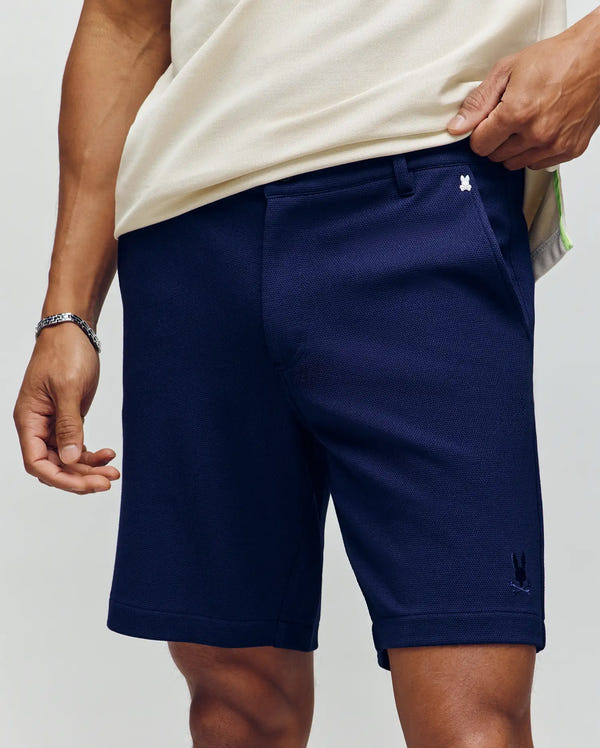 Wearing Psycho Bunny's MENS SHIRO KNIT HONEYCOMB SHORT in dark navy with pockets, detailed in a honeycomb-patterned piqué and an embroidered Bunny logo near the waistband, they pair it with a light shirt and a silver bracelet.