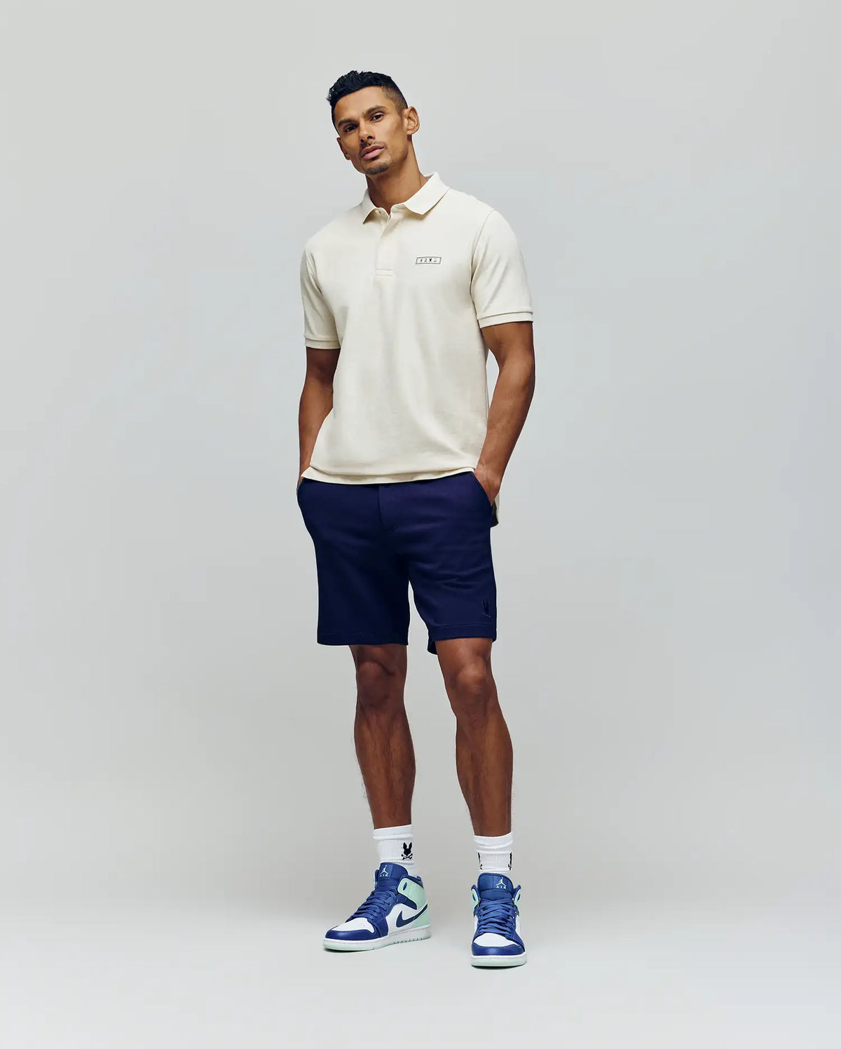 A man poses confidently against a light gray background wearing a cream polo, Psycho Bunny MENS SHIRO KNIT HONEYCOMB SHORTS, white socks, and blue-and-white sneakers. An embroidered Bunny logo adds a playful touch to his look.