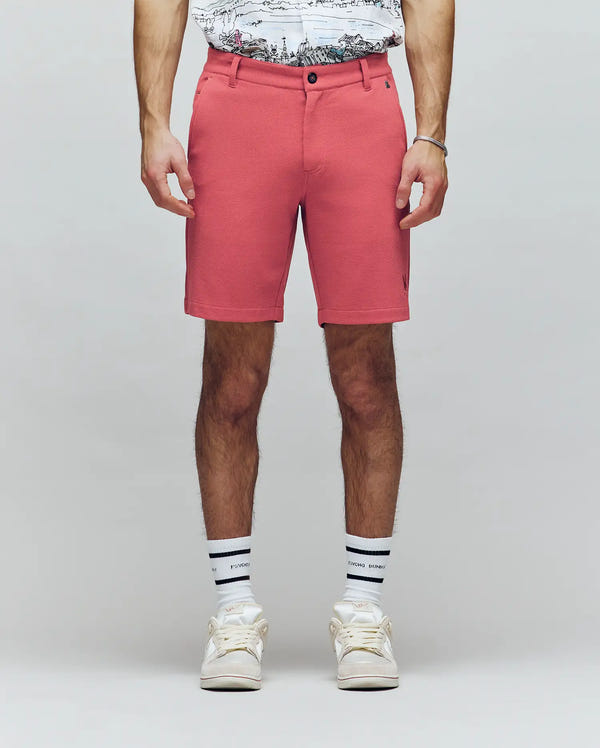 A person stands against a plain light background, shown waist to feet, wearing Psycho Bunny MENS SHIRO KNIT HONEYCOMB SHORT - B6R585F200 in coral pink, a white printed t-shirt, white socks with black stripes, and white sneakers.