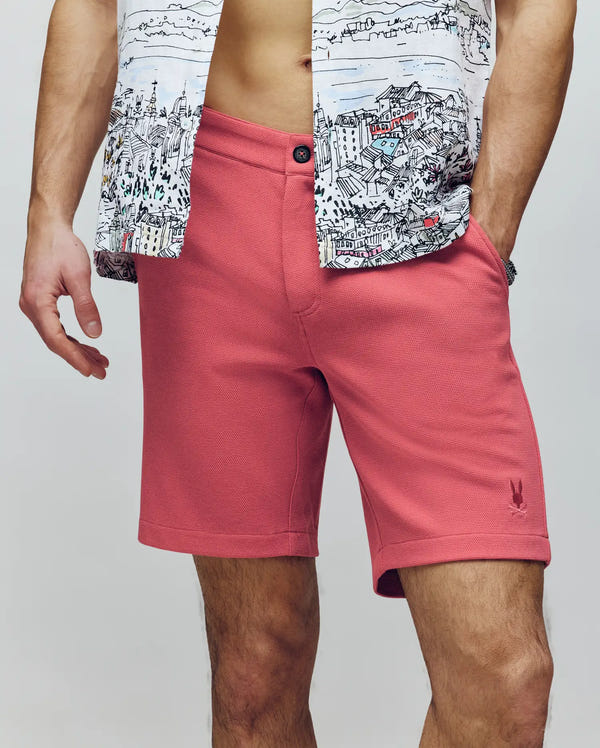 A person in Psycho Bunny's MENS SHIRO KNIT HONEYCOMB SHORT - B6R585F200 (salmon) and a cityscape-print unbuttoned shirt, torso exposed. An embroidered Bunny accent adds subtle charm; only their lower face and body appear.
