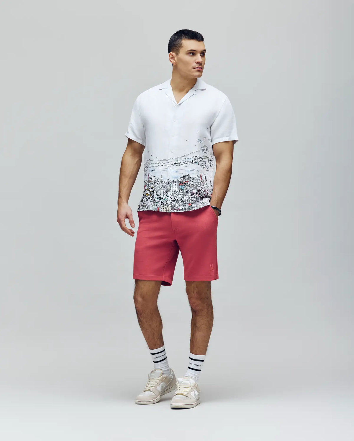 A man stands against a plain background in a white short-sleeve shirt with an embroidered Bunny cityscape, Psycho Bunny MENS SHIRO KNIT HONEYCOMB SHORT - B6R585F200 in pink, white socks with black stripes, and white sneakers.