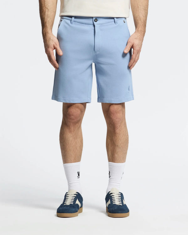 A person is wearing Psycho Bunny's MENS SHIRO KNIT HONEYCOMB SHORT - B6R585F200 in light blue with a honeycomb pattern, white socks, and dark blue sneakers. Standing against a plain background, both hands are in their pockets, showcasing the shorts' embroidered bunny logo on the left.