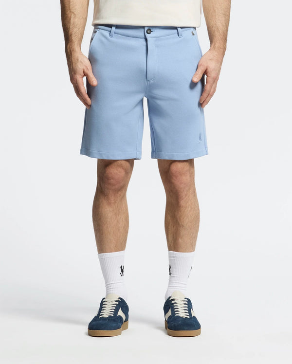 A person is wearing Psycho Bunny's MENS SHIRO KNIT HONEYCOMB SHORT - B6R585F200 in light blue with a honeycomb pattern, white socks, and dark blue sneakers. Standing against a plain background, both hands are in their pockets, showcasing the shorts' embroidered bunny logo on the left.