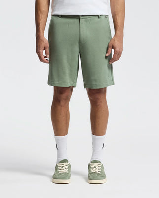 A person wears the Psycho Bunny MENS SHIRO KNIT HONEYCOMB SHORT - B6R585F200, paired with piqué sneakers and crisp white socks against a plain white background, showcasing just their lower half.