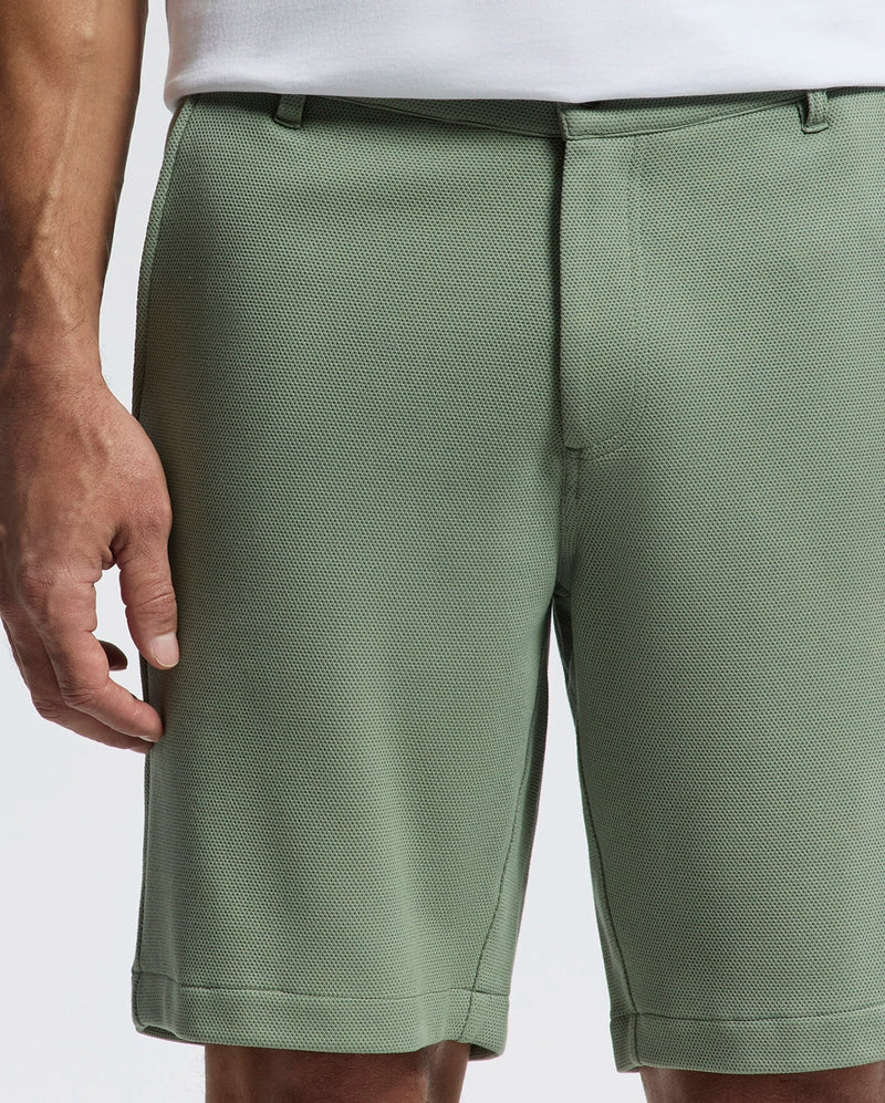 A person is shown from the waist down wearing Psycho Bunny's MENS SHIRO KNIT HONEYCOMB SHORT - B6R585F200 in light green piqué. They are also in a white shirt, with their left arm partially visible against a plain white background.