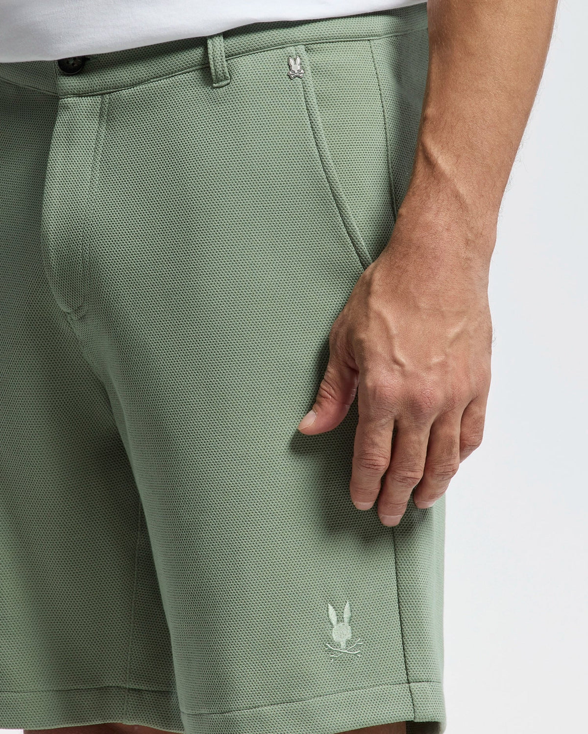 A person is wearing light green Psycho Bunny's MENS SHIRO KNIT HONEYCOMB SHORT - B6R585F200, featuring a small bunny logo, honeycomb piqué texture, visible pockets, and belt loops. Their hand is next to the pocket against a plain white background.