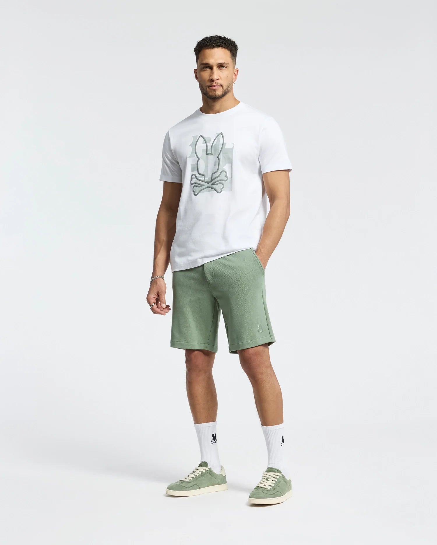 A man poses against a plain, light background in a white graphic T-shirt, versatile green MENS SHIRO KNIT HONEYCOMB SHORT by Psycho Bunny, matching green sneakers, and white socks with black details.
