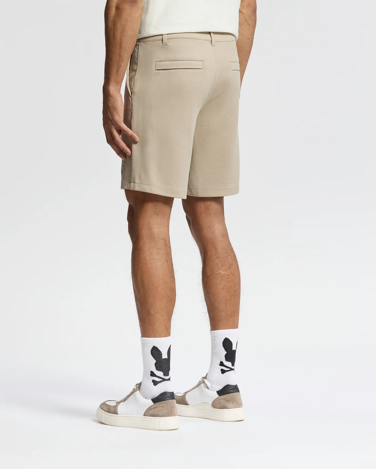 A man, back turned, wears Psycho Bunny MENS SHIRO KNIT HONEYCOMB SHORTS (B6R585F200), white socks with black graphics, white sneakers with tan accents, and a light shirt against a plain white background for a smart-casual look.