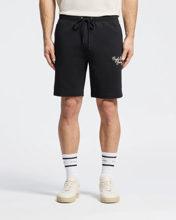 The Psycho Bunny MENS DESERT EMBROIDERED MODAL SWEATSHORT - B6R617F200 in black is styled with a crisp white shirt, complemented by white crew socks with bold black stripes and sleek white sneakers, all against a plain background.