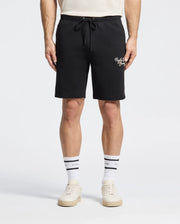 The Psycho Bunny MENS DESERT EMBROIDERED MODAL SWEATSHORT - B6R617F200 in black is styled with a crisp white shirt, complemented by white crew socks with bold black stripes and sleek white sneakers, all against a plain background.