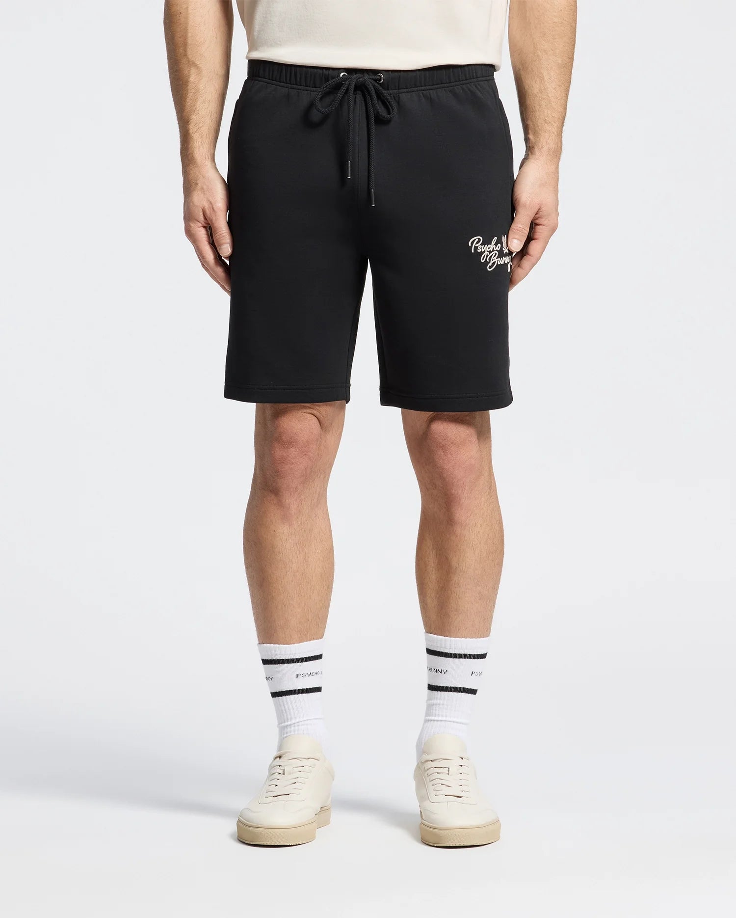 The Psycho Bunny MENS DESERT EMBROIDERED MODAL SWEATSHORT - B6R617F200 in black is styled with a crisp white shirt, complemented by white crew socks with bold black stripes and sleek white sneakers, all against a plain background.