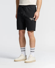 The person is wearing Psycho Bunny's Men's Desert Embroidered Modal Sweatshorts (B6R617F200) in black, featuring white text and an embroidered graphic by the pocket, paired with a beige T-shirt, white crew socks with black stripes, and white sneakers against a plain white background.