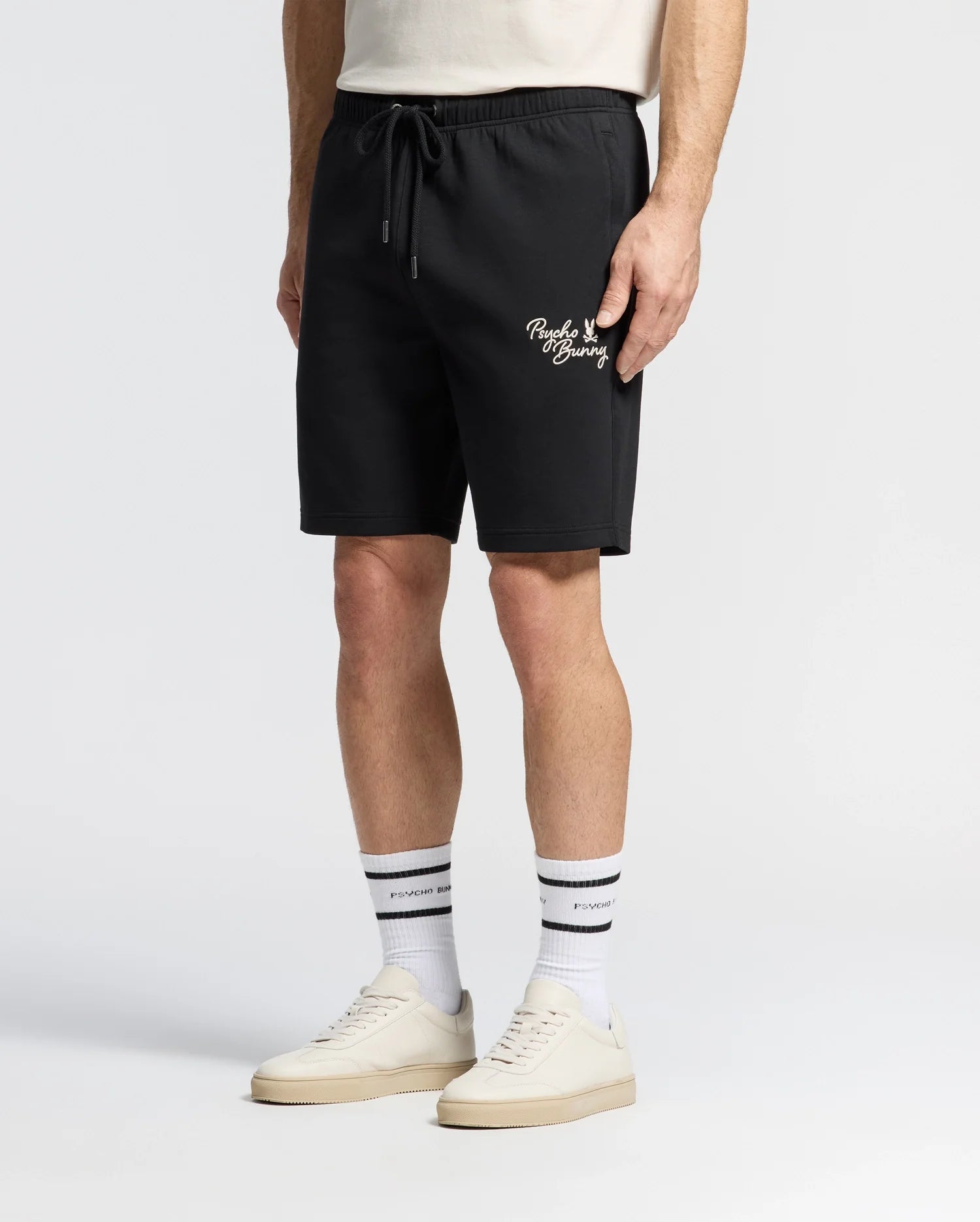 The person is wearing Psycho Bunny's Men's Desert Embroidered Modal Sweatshorts (B6R617F200) in black, featuring white text and an embroidered graphic by the pocket, paired with a beige T-shirt, white crew socks with black stripes, and white sneakers against a plain white background.