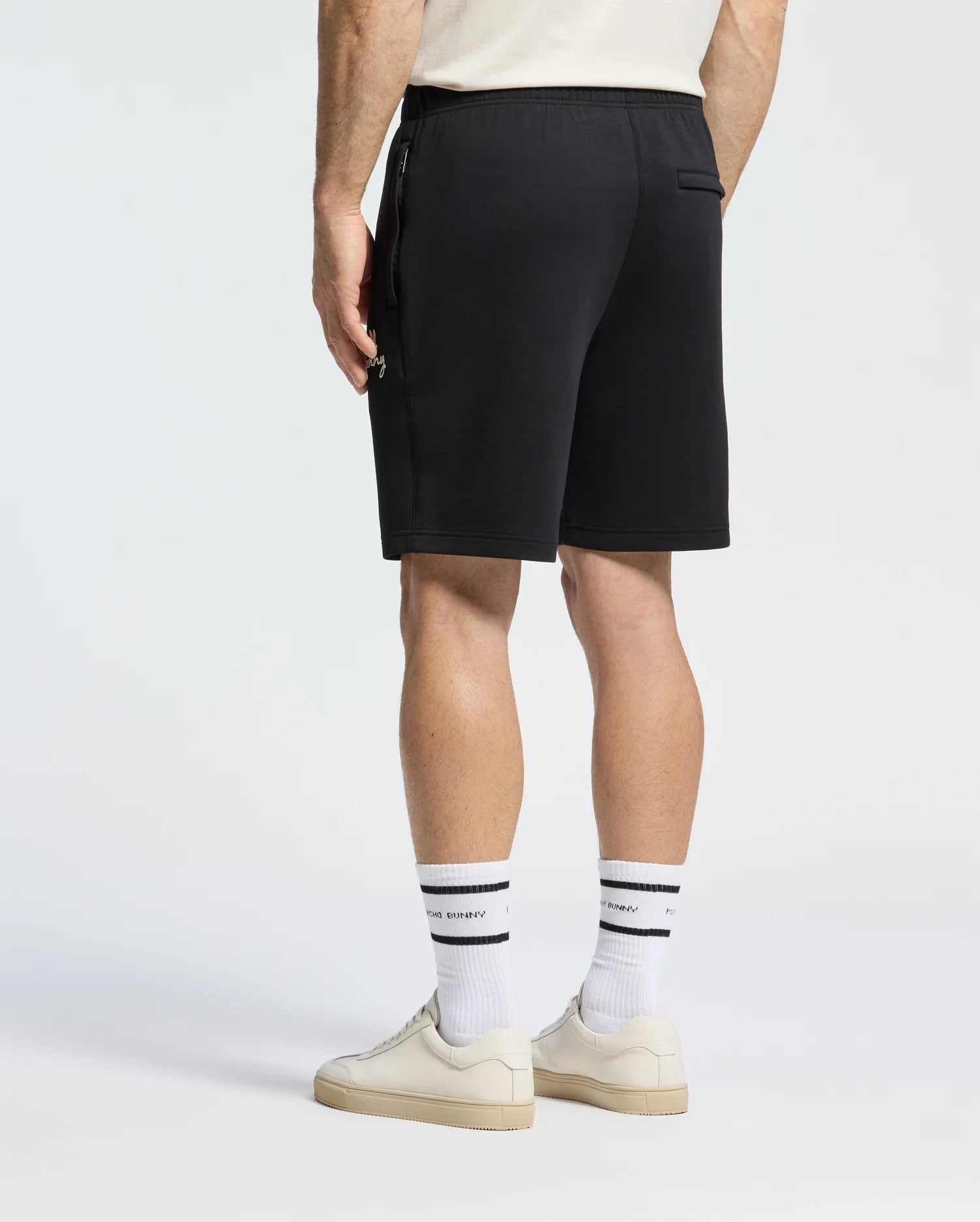 Someone in Psycho Bunny's MENS DESERT EMBROIDERED MODAL SWEATSHORTS, a white shirt, and white socks with black stripes stands sideways against a grey background. Their face is not visible. They are also wearing white sneakers.