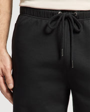 A person is seen wearing Psycho Bunny's black MENS DESERT EMBROIDERED MODAL SWEATSHORT (B6R617F200), featuring an embroidered graphic and a drawstring waist. Their left hand rests by their side, complementing their light shirt.
