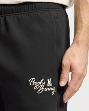 A person wearing black Psycho Bunny men's desert embroidered modal sweatshorts with a small bunny logo in moisture-wicking fabric stands with one hand on their leg.