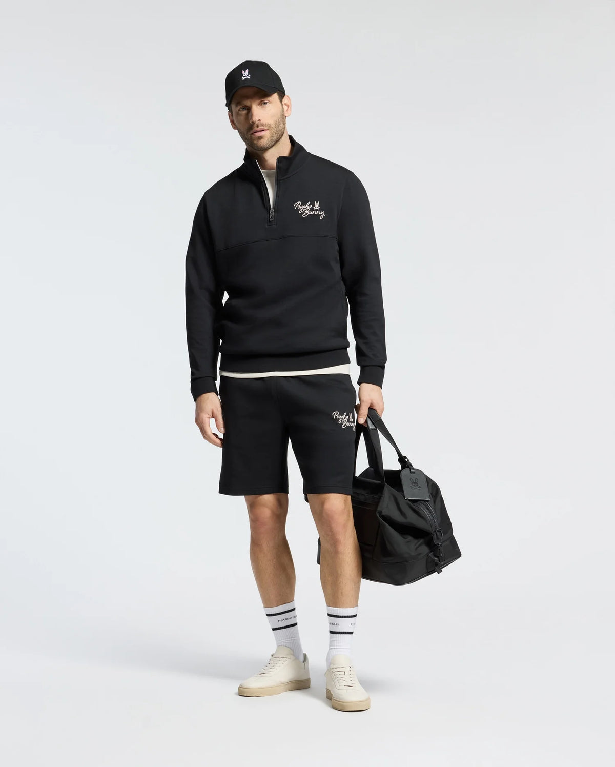 A man, wearing a black cap, quarter-zip sweatshirt, and Psycho Bunny's MENS DESERT EMBROIDERED MODAL SWEATSHORT - B6R617F200, stands with a black duffel bag. He dons white socks and beige shoes against a plain backdrop. The outfit includes subtle embroidered graphics for a casual sporty vibe.