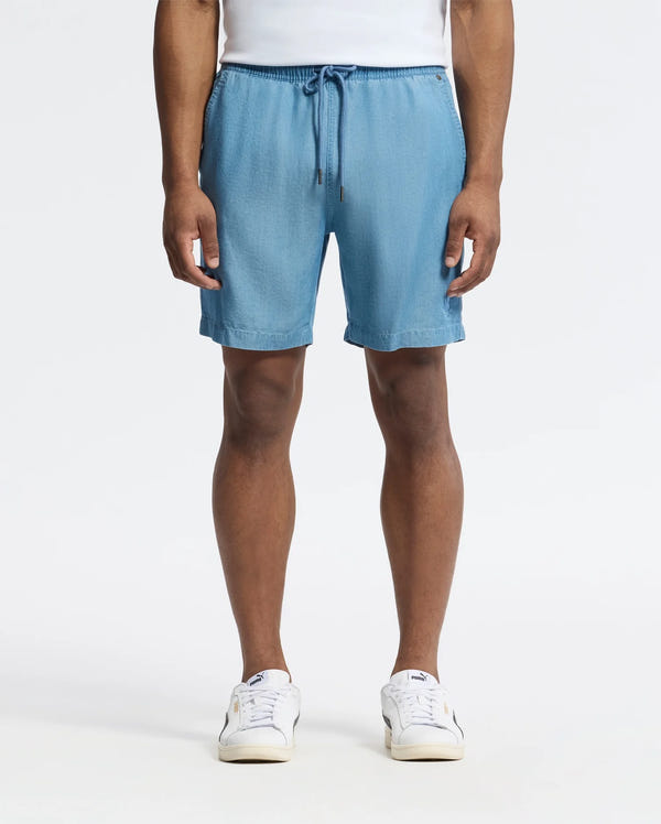 A person models Psycho Bunny's MENS KERVIN LIGHTWEIGHT DENIM WOVEN SHORT (B6R633F200) in light blue, featuring an embroidered bunny and elastic waist, paired with white sneakers against a plain white background. Only their lower half is visible, hands relaxed at their sides.