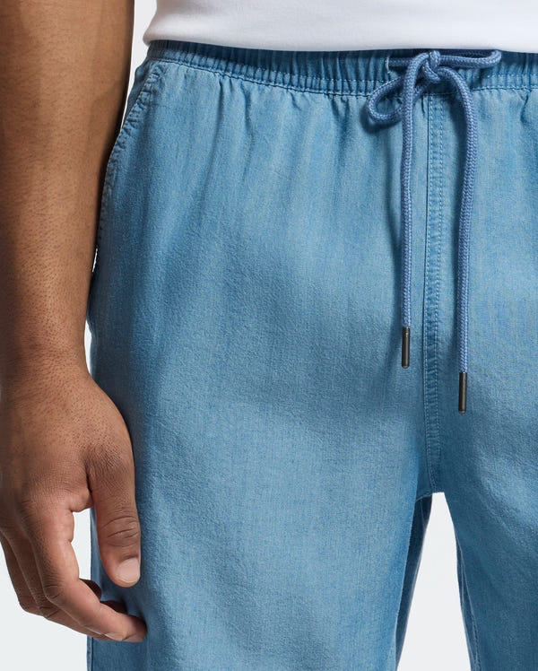 A close-up of a person wearing Psycho Bunny's Mens Kervin Lightweight Denim Woven Short in light blue, featuring an elasticized waist and relaxed fit, paired with a white shirt. The shorts are showcased from the waist to the knee with the person's hand partially visible on the left side.