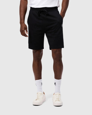 A person is wearing Psycho Bunny's MENS LEVY FLEECE SWEATSHORT - B6R862D200 in black, regular fit with a drawstring waist, paired with white socks and white sneakers. The French terry shorts feature a small logo on the left leg. The background is plain light gray.