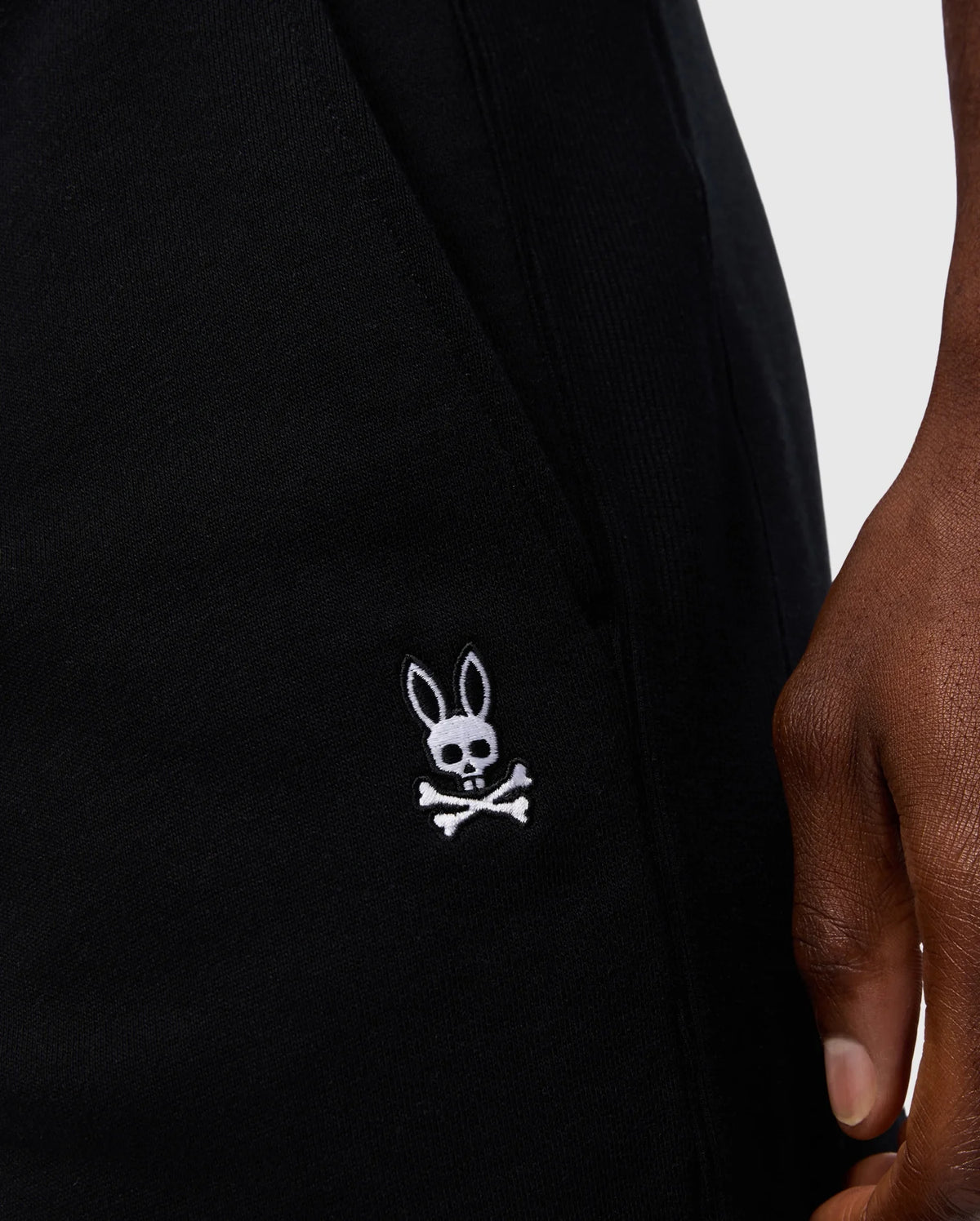 Close-up of a person's hand beside the pocket of Psycho Bunny's MENS LEVY FLEECE SWEATSHORT - B6R862D200, featuring an embroidered logo of a cartoon skull and crossbones with bunny ears. The background is light gray.