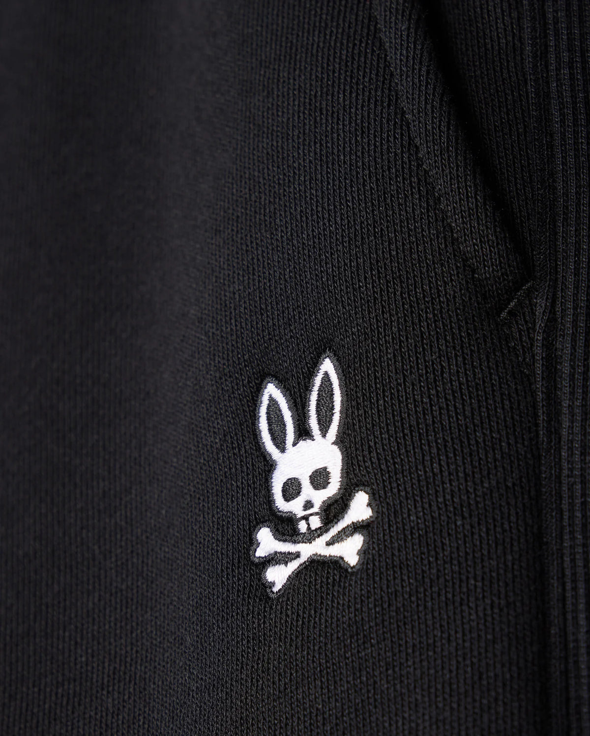 Close-up of the Psycho Bunny MENS LEVY FLEECE SWEATSHORT - B6R862D200, a black cotton fleece sweatshort with an embroidered white logo featuring a rabbit head with crossbones underneath.