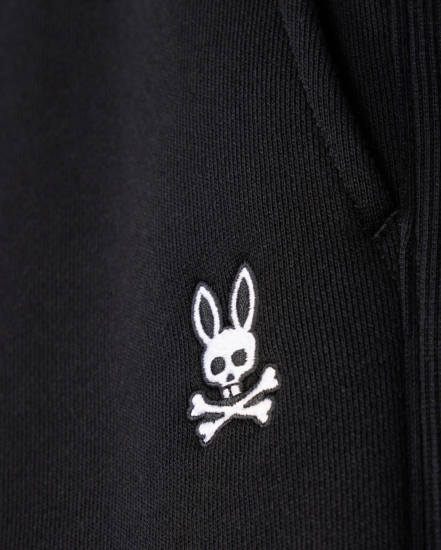 Close-up of the Psycho Bunny MENS LEVY FLEECE SWEATSHORT - B6R862D200, a black cotton fleece sweatshort with an embroidered white logo featuring a rabbit head with crossbones underneath.