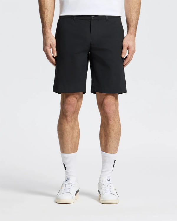 A person wearing Psycho Bunny's MENS 10K STEP SHORT - B6R969F200, along with white crew socks and sneakers, stands against a plain backdrop. The image features only their legs and part of the torso.
