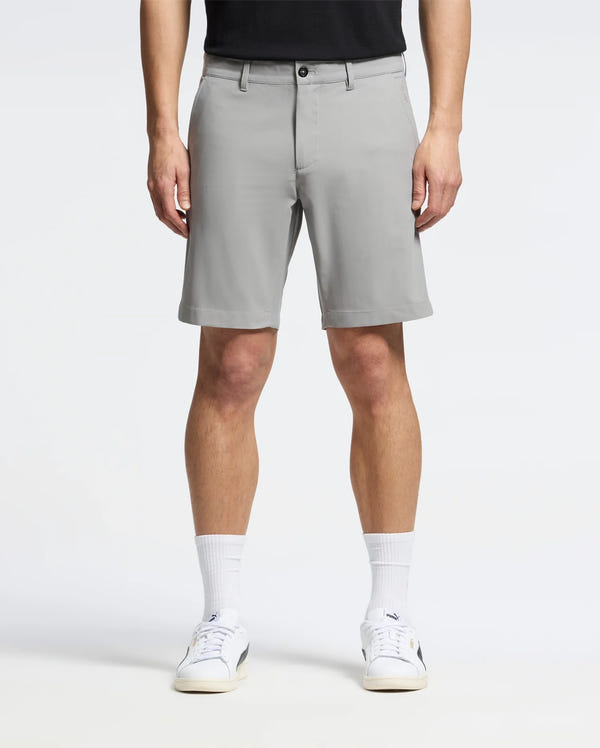 A person wearing Psycho Bunny MENS 10K STEP SHORT - B6R969F200 in light grey pairs them with a black shirt, white socks, and sneakers against a plain backdrop. The shorts have belt loops and button closure for style and comfort.