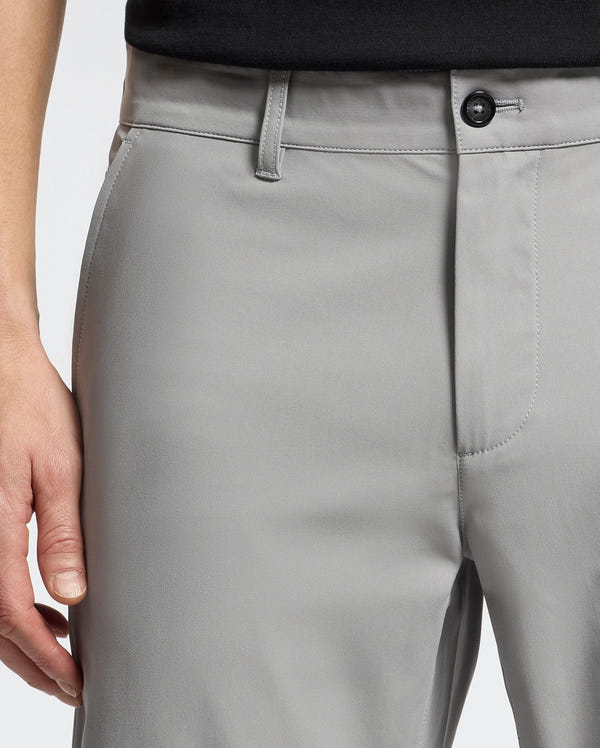 A close-up of a person donning Psycho Bunny's MENS 10K STEP SHORT - B6R969F200. The light gray shorts offer athletic breathability, a button, zipper, and belt loops. A black shirt complements the style as their left hand rests at their side, highlighting comfort and flair.
